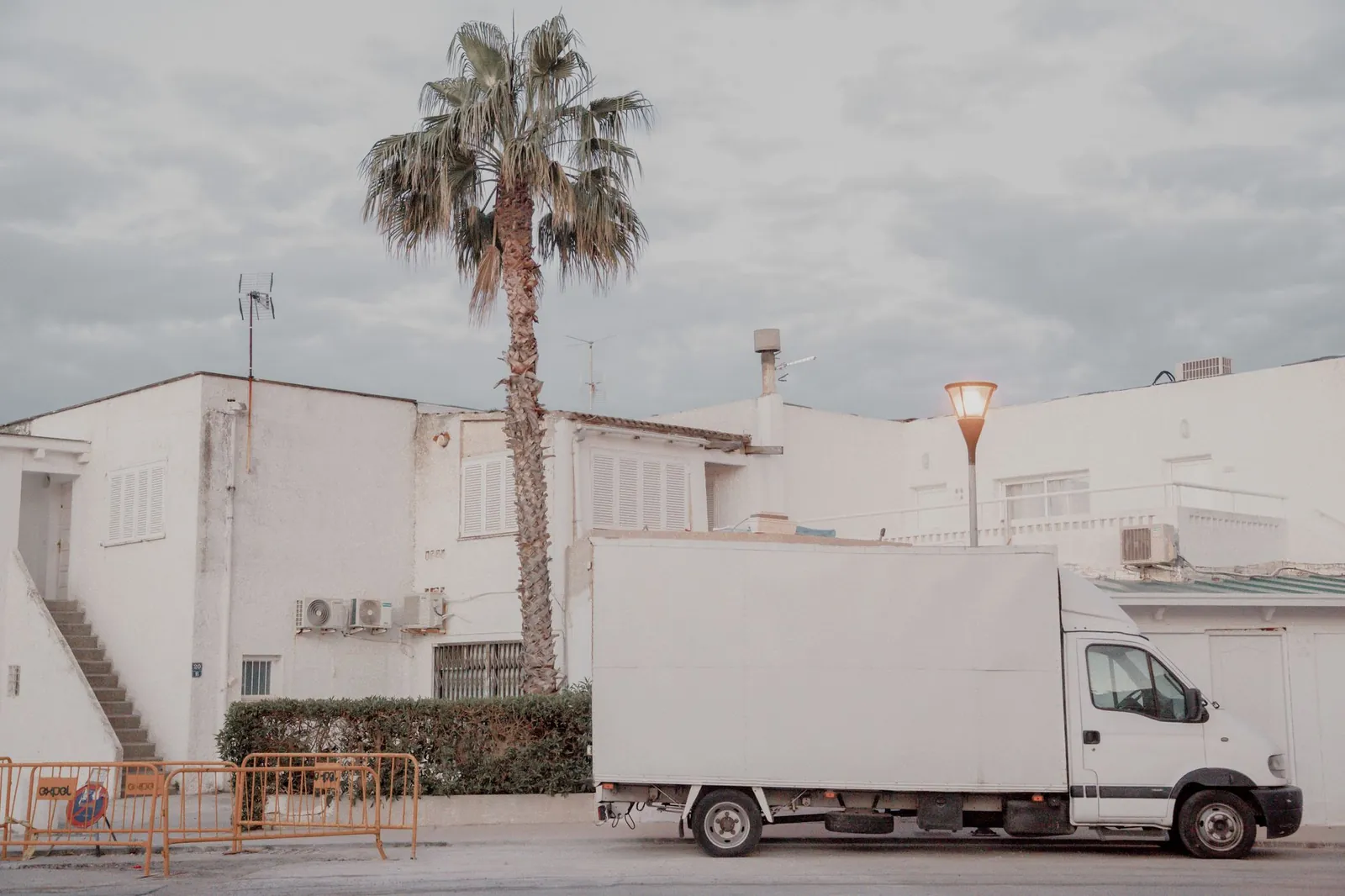 Plain, unbranded white box truck parked on a street in front of white stucco Mediterranean-style buildings with a tall palm tree, overcast p