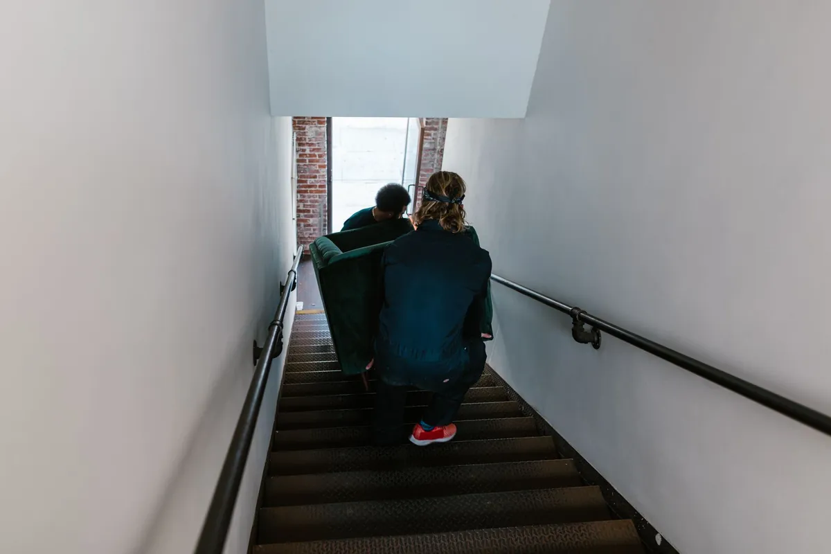 Two movers carrying a green sofa down a narrow apartment stairwell with black handrail, seen from behind/above